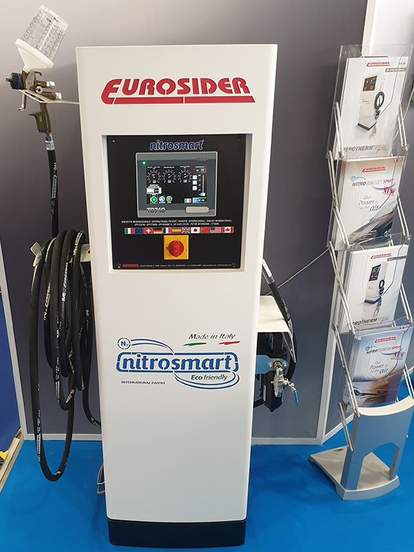 Nitrosmart framifrån ståendes på en blå matta. Maskinen har en stor display i mitten och på vänster sida hänger en svart slang med ett munstycke längst upp.
                                Ett ställ med broschyrer står på höger sida.