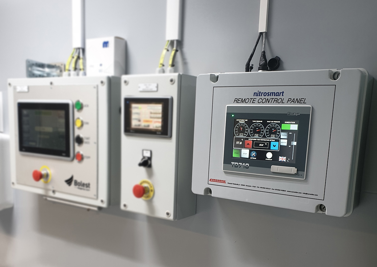 Närbild på kontrollpanel placerad på väggen. Panelen är grå och har texten Nitrosmart Remote Control Panel. Ofokuserat i bakgrunden finns fler displayer.